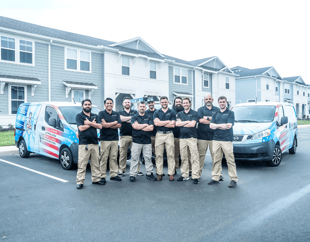 A row of Air Titans HVAC professionals in black polos and khaki pants stand with arms crossed and smile in front of two company vans parked outside a modern apartment complex.