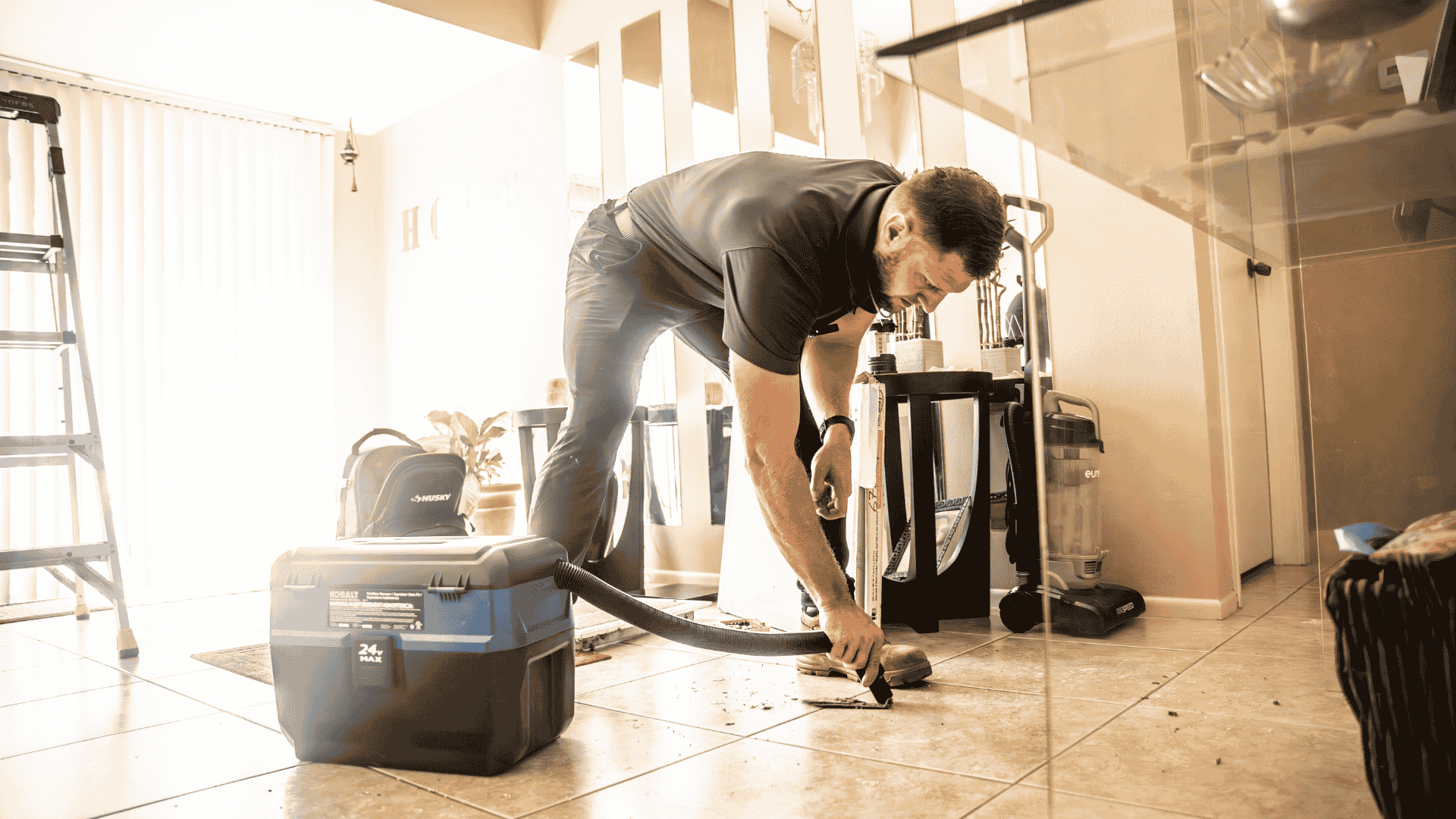 Technician bends to vacuum debris from under a dining table near the HVAC return, preparing the home’s air conditioning system for optimal airflow.