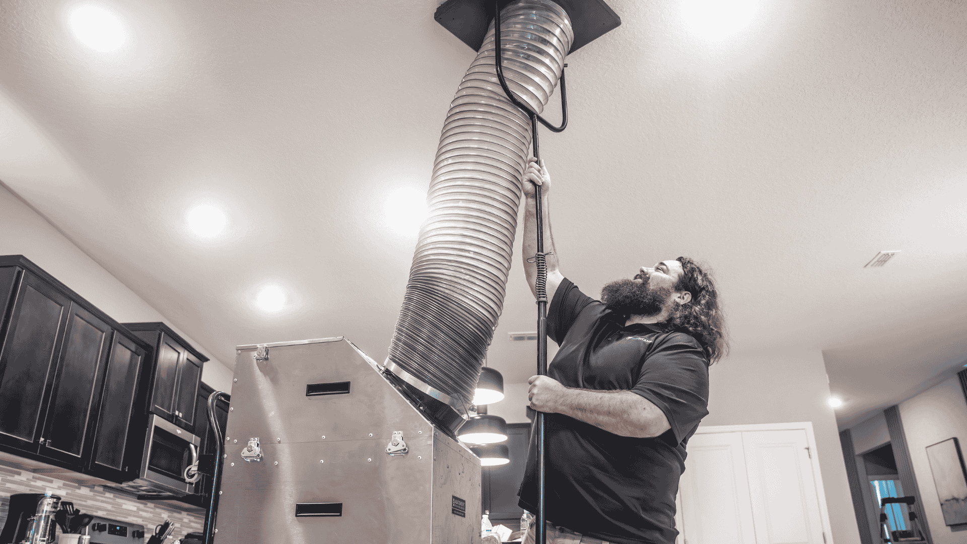 Technician feeds a large flexible hose into a ceiling dryer‐vent opening above a stainless steel duct head, clearing lint and buildup.